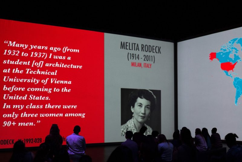 An audience views two walls of screens, one showing a map and another a headshot and quote from architect Melita Rodeck. 