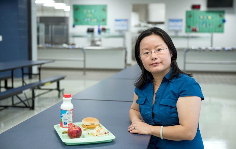 A woman sits in a school cafeteria in front of a cafeteria tray of food.a
