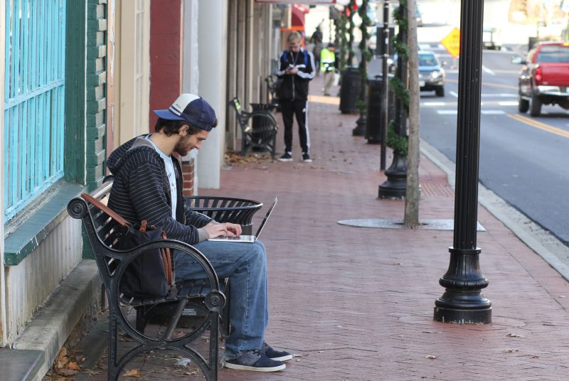 student on bench along Main Street in Blacksburg, working on a laptop