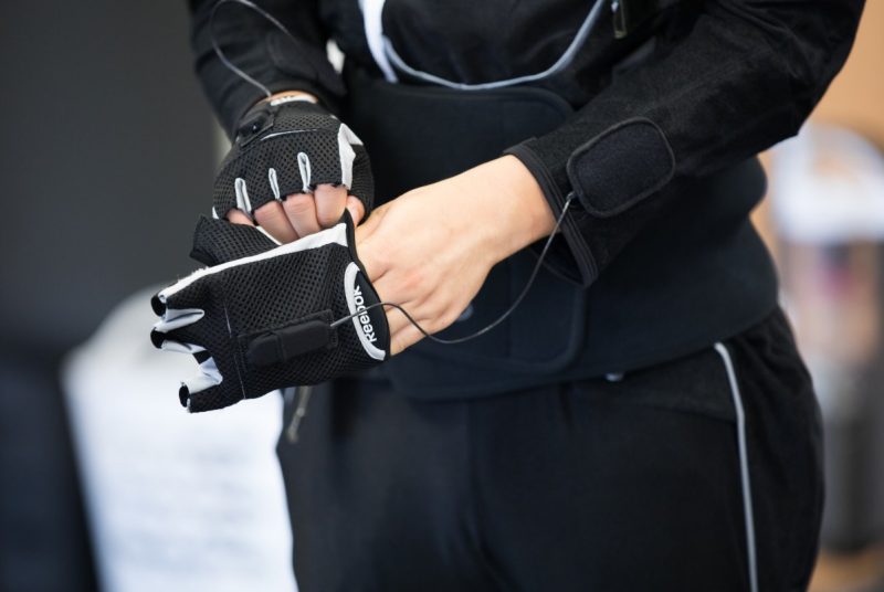 In the photo, a young man tries on a black cloth glove, which is connected to wires. It's a close up shot, focused only on the lower torso and the black suit.