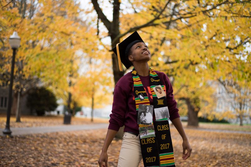 Graduating senior Monica Fikes in commencement gear amidst autumn leaves.