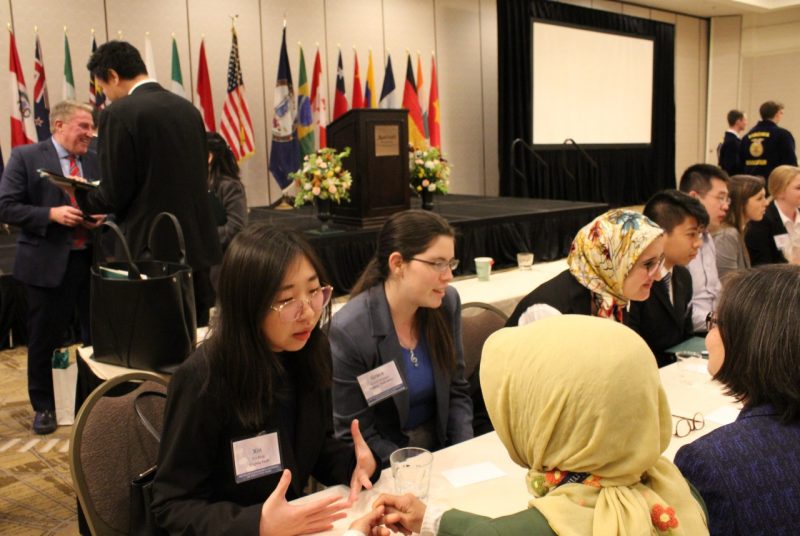 Students sit at table discussing trade issues with international trade representatives. Other conference attendees network in the background.
