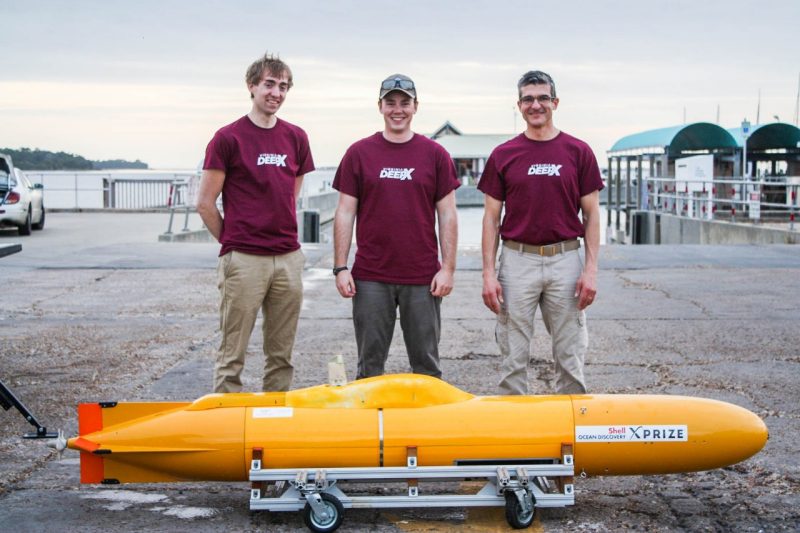 Three men pose with a small, orange autonomous vehicle that looks like a small submarine.