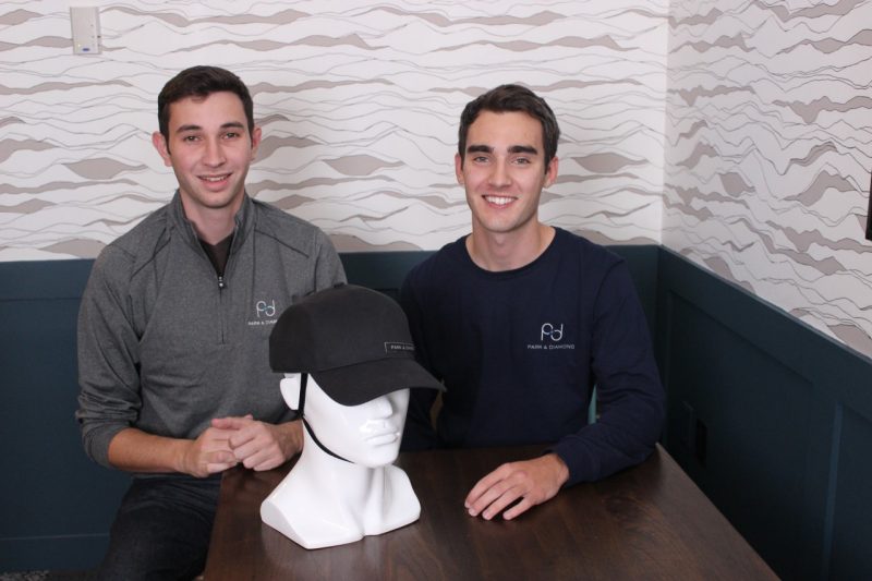Two young men sit at a table and pose for a photo. On the table is a white mannequin head and neck, wearing a black helmet that looks like a baseball cap.