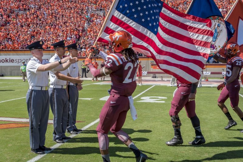 Flag cadets Cadet John Mitchell, at left, reaches for the American flag carried by a Hokie football player. To his right are Cadets Abdel Hammad and Paul Pechie.