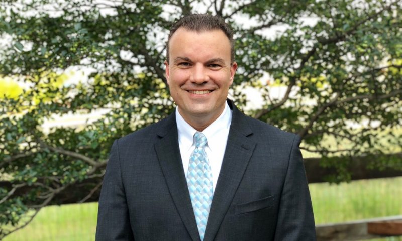 An image of Ryan McDaniel wearing a suit and tie, smiling, and standing outdoors in front of a tree.