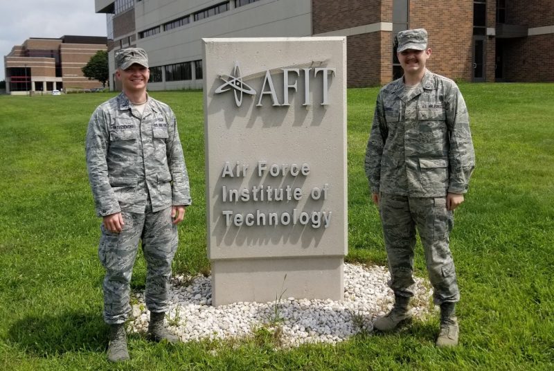 Two men in military fatigues standing on either side of a sign with a building in the background.