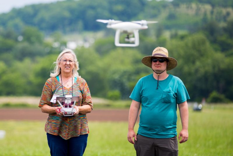 A woman stands in an open area holding a controller, a man stands next to her. A small white drone hovers in front of them.