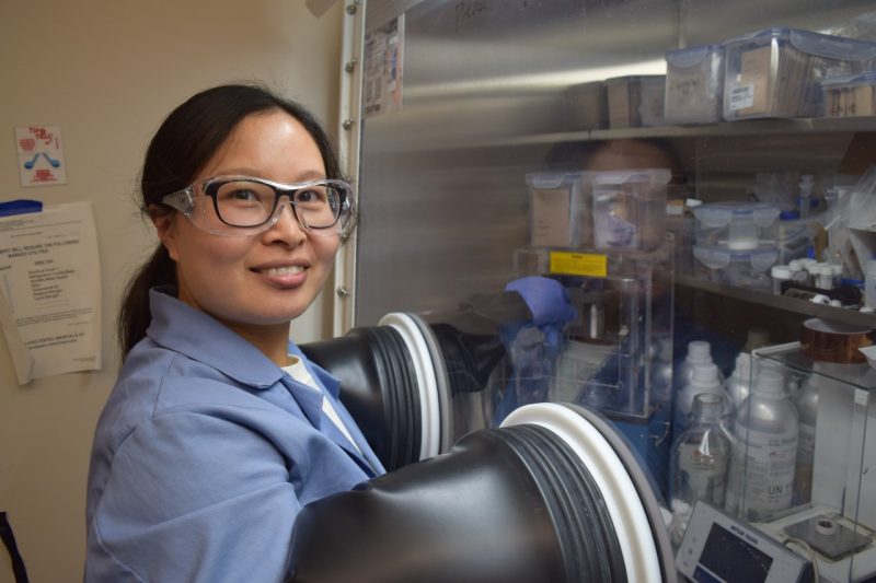 Linqin Mu, wearing safety glasses and a blue lab coat, smiles while working in the glove box.