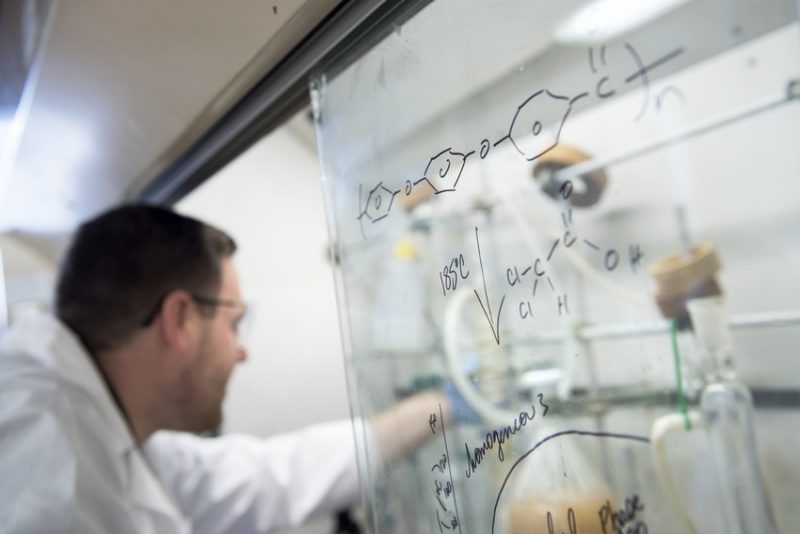 A science researcher works under the hood in the background. A science equation is written on a board in the foreground.