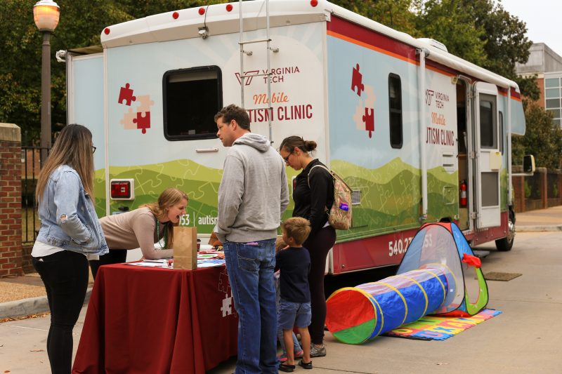 Two parents arrive with their child to a booth that is adjacent to the Mobile Autism Clinic van, which is adorned with mountains, maroon puzzle pieces, and the Virginia Tech logo. Two members from the Center for Autism Research greet the child with excitation.