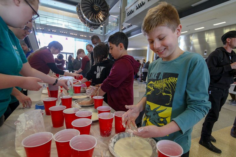 A child is smiling as he observes his hands after creating a beige-colored slime in a foil pan. A few red solo cups are surrounding the slime station that is on the table.