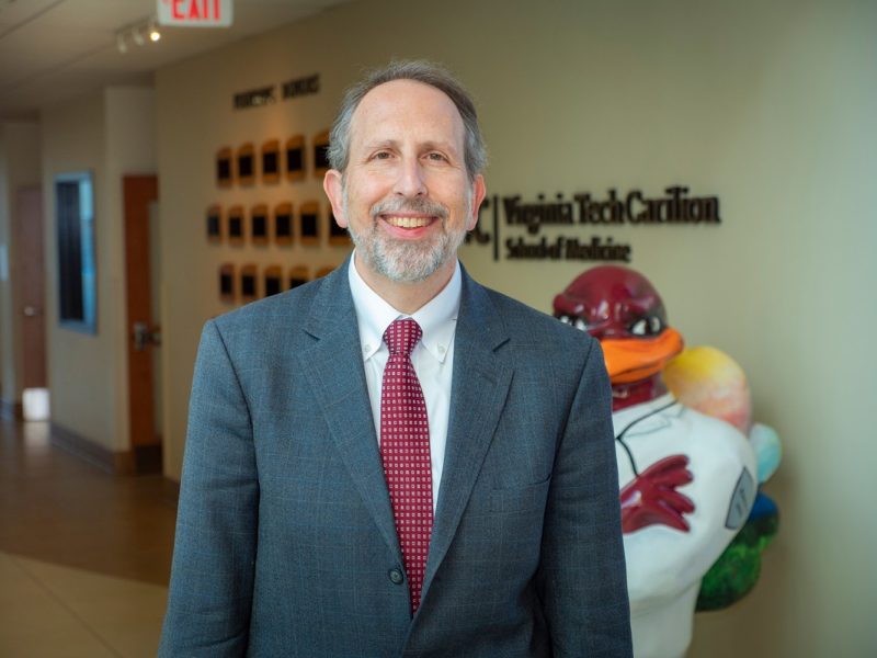 Lee Learman stands in the medical school atrium just in front of a logo on the wall and a HokieBird