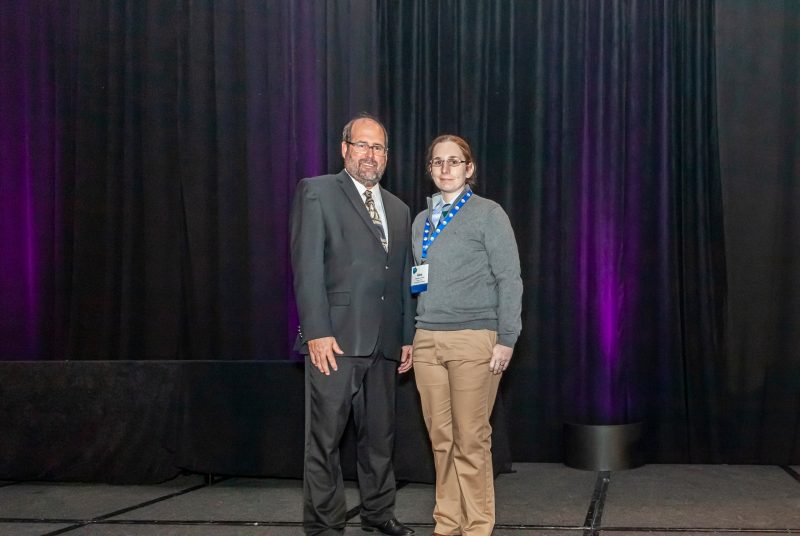 A man is presenting Alexandria Noble, a graduate student at Virginia Tech, with an award. They are standing on a stage with a purple curtain behind them.