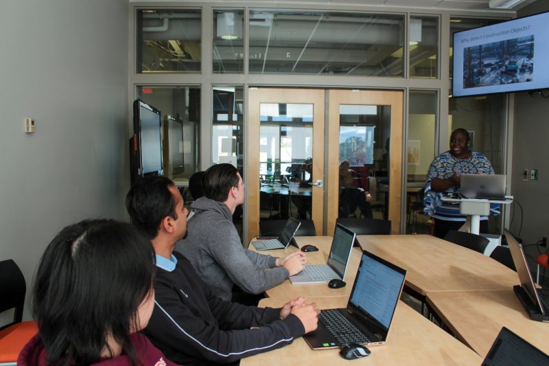 Students with laptops in classroom listen to Dr. Akanmu lecturing in front of computer screen. 