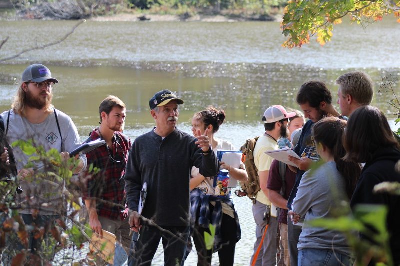A Virginia Tech professor speaks to students with his finger pointed. They are all working in the field near a creek.