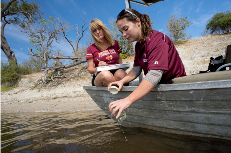 Two women in Virginia Tech shirts are sitting in a boat that lies on a bank of the Chobe River in Botswana. The young graduate student is collecting a jar of water off of the side of the boat, as her professor sits behind her with a large clipboard and a pen.