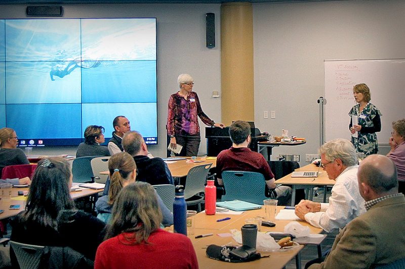 Two women are standing and speaking to a crowded group of tables. A large screen is behind one woman and a whiteboard is behind the other. Tables are adorned with food, water bottles, and laptops.