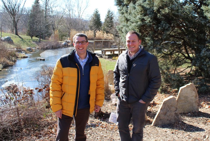 Two men stand outdoors with a stream and trees behind them.