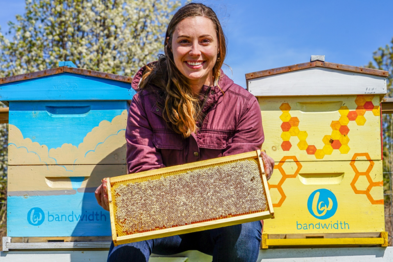 Bee Downtown founder and CEO Leigh-Kathryn Bonner, who is holding up a frame that is covered in honeycombs. She is standing in between two brightly painted beehive boxes, one depicting a partly cloudy sky, and the other depicting honeycombs. Courtesy: Bee Downtown.