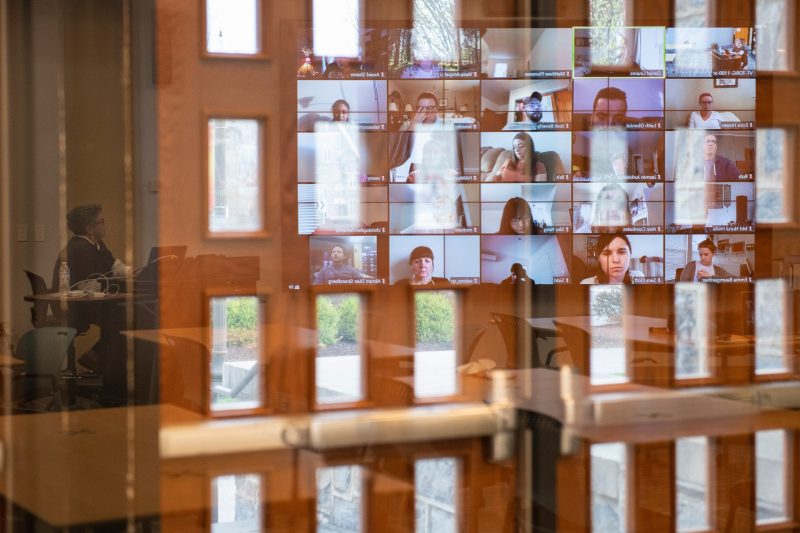 Photo shows reflections of various student faces on their individual screen monitors as they meet virtually for a class taught by Karen DePauw, shown seated at a desk in the empty classroom