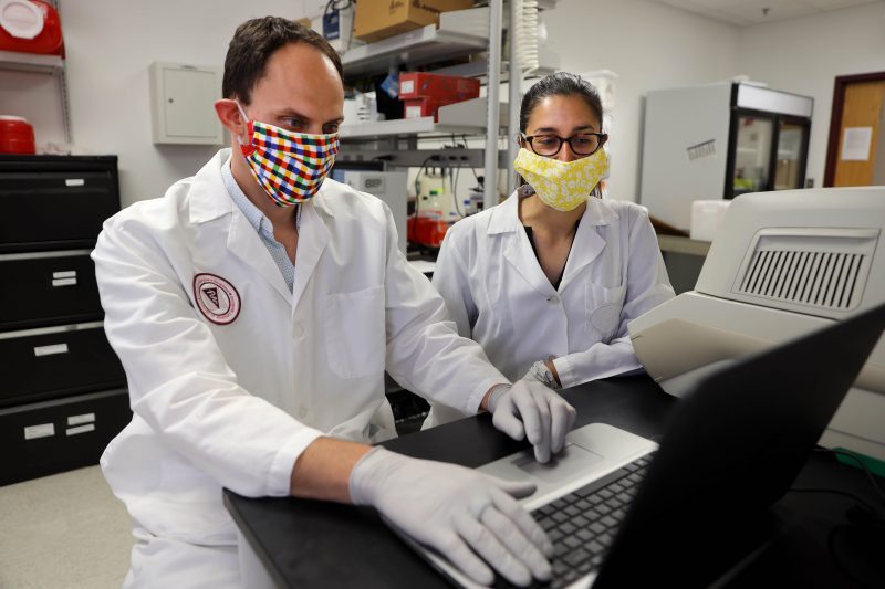 James Weger Lucarelli (left) and Nisha Duggal (right) conducting research in the lab. Lucarelli is wearing a rainbow colored mask while using a laptop. Duggal watches on with a yellow face mask. Ray Meese for Virginia Tech.