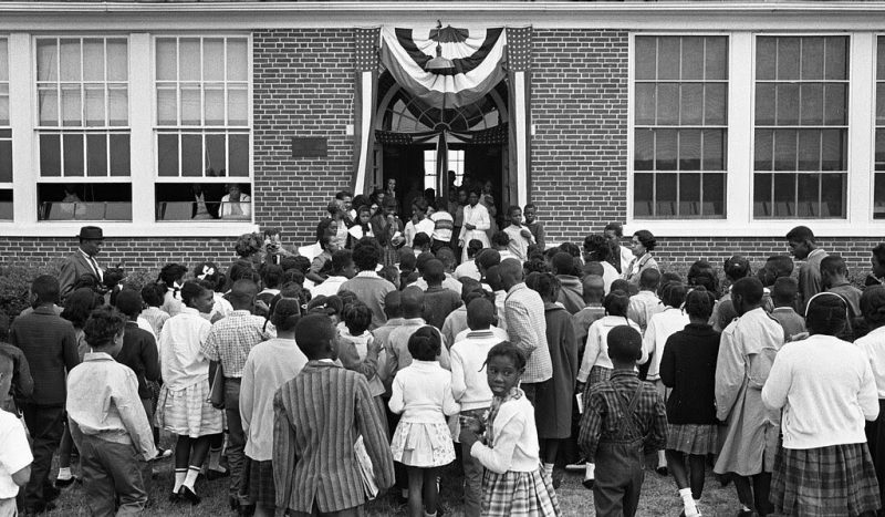  Dozens of young black children enter the double doors of a school. One black man in a hat and several black women in glasses enter with them. The front door is decorated with American flags and streamers. Two people watch the crowd from inside through the windows. “African American school children entering the Mary E. Branch School at S. Main Street and Griffin Boulevard, Farmville, Prince Edward County, Virginia.” O'Halloran, Thomas J, photographer. African American school children entering the Mary E. Branch School at S. Main Street and Griffin Boulevard, Farmville, Prince Edward County, Virginia. Farmville Virginia, 1963. Sept. 16. Photograph. https://www.loc.gov/item/2011648793/. No known restrictions on publication.
