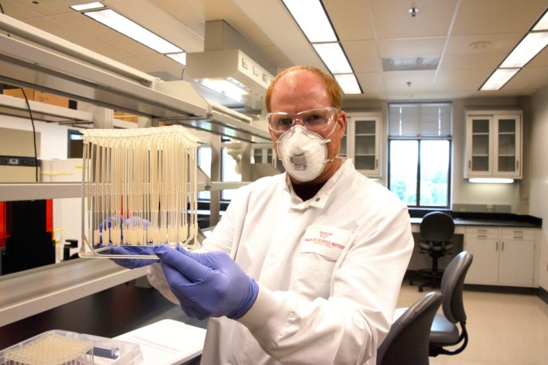 Marc Michel, donning PPE mask, lab coat, and gloves, holds rack of Covid-129 test swabs.