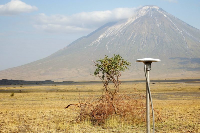 A tall GPS positioning sensor stands at the base of the massive Ol Doinyo Lengai (The Mountain of God), an active volcano in Tanzania. The volcano looms large in the distance. 