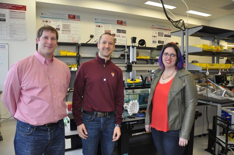 Irving Coy Allen (left), Eli Vlaisavljevich (center), and Jennifer Munson (right) stand in a biomedical engineering and mechanics research lab. A method used in this research is a technique called histotripsy. The device used for this technique is pictured in the background. Photo credit Laura McWhinney of Virginia Tech. [Note: Picture taken before the COVID-19 pandemic].