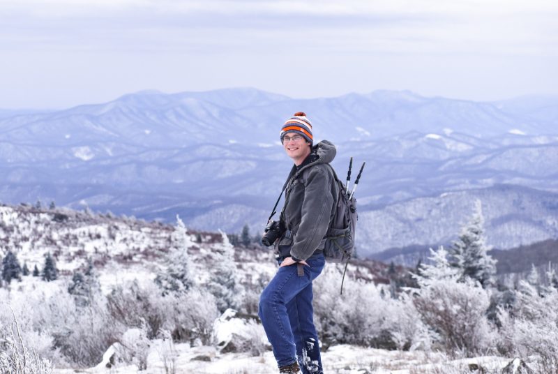 A man with a backpack and camera stands on a snowy mountaintop.