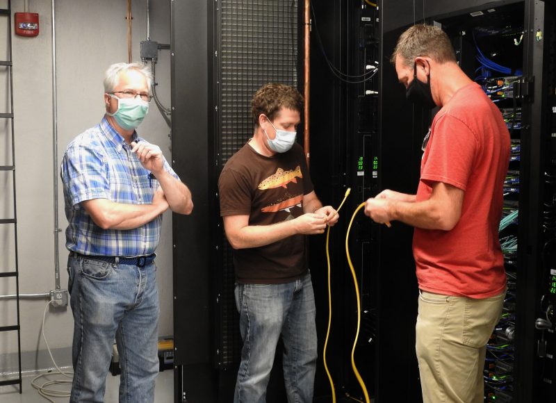 three systems engineers wearing face coverings work on supercomputing hardware in data center