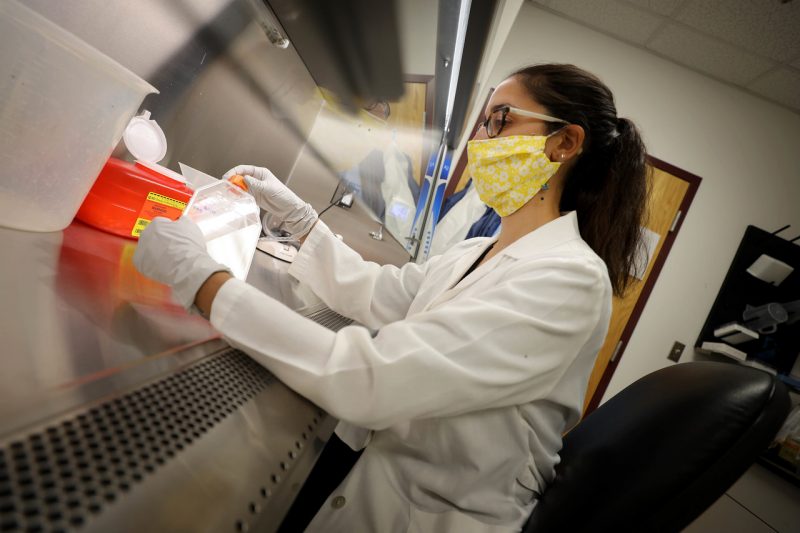 A researcher wearing a mask and a lab coat sitting at a lab bench