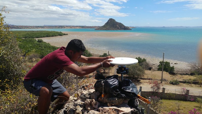 Geosciences doctoral student Tahiry Rajaonarison sets up a GPS instrument in northern Madagascar in this 2016 photograph. Behind Tahir is the Indian Ocean and a rock island.  Photo credit: Rina Andrianasolo.