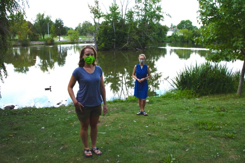 Nicole Ward, a doctoral student, and Cayelan Carey, an associate professor, both in the Department of Biological Sciences pose at the Duck Pond. Each is wearing a mask and standing more than six feet part.
