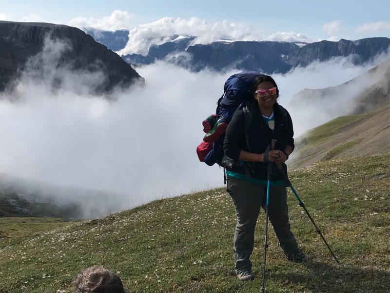 Ph.D. student Selva Marroquín backpacking to the field site after being dropped off by a bush plane in Wrangell-St. Elias National Park, Alaska. Behind Marraquin is a vat mountain range with clouds.