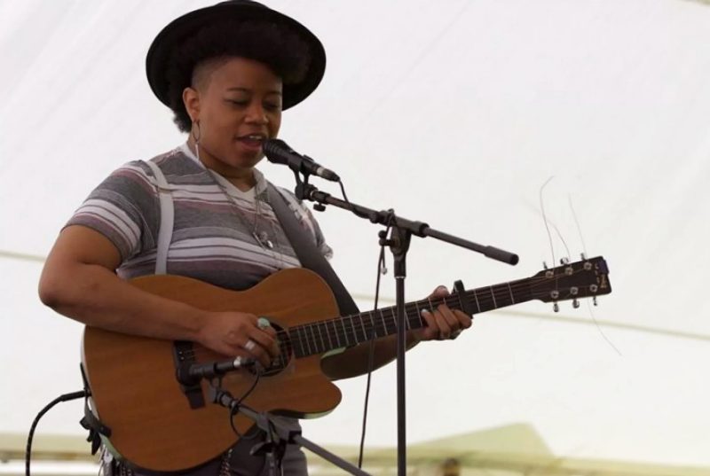 A young black performer plays a guitar and stands at a microphone sining.
