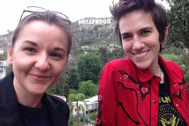 Two women  with the Hollywood hills sign behind them; one cearing a black shirt with classes on her head, the other wearing a red western style shirt.