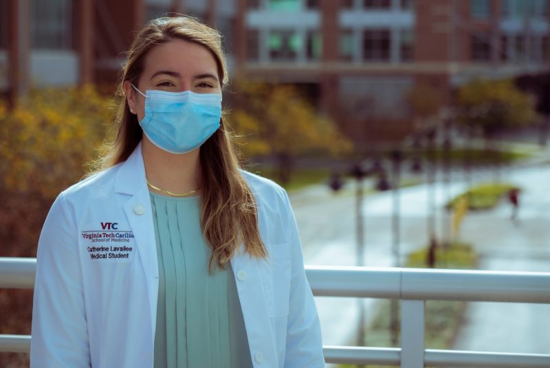 Young femaile wearing doctor's white coat and mask standing outdoors with buildings in background.