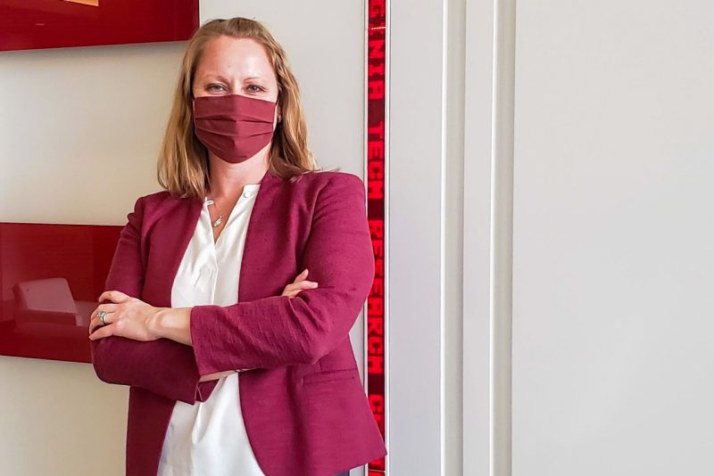A blonde woman in a maroon mask and business coat stands in front of a white wall.