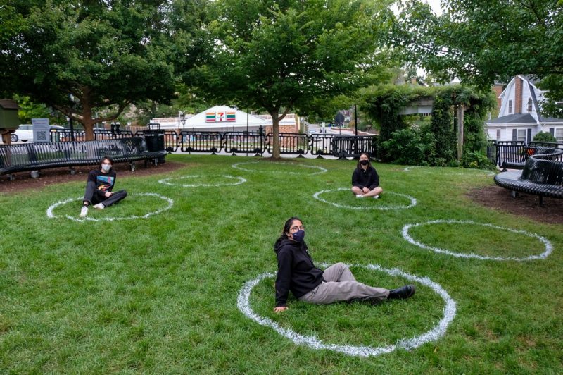 Jake Swartz (left), Dorsa Naimi (center), and Joonyoung Kang (right) sit inside the circles spray painted on the ground as part of the Hokie Circles project to encourage physical distancing at the Blacksburg Farmers Market. Photo by Aki Ishida.
