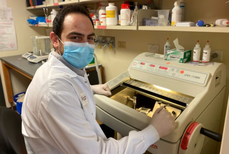 Man wearing white lab coat sitting at lab equipment.