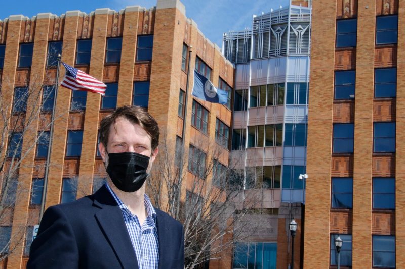 David Conners, wearing a black face mask, stands in front a tall brick building.