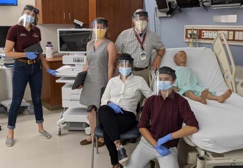 Three members of the transdisciplinary research team are standing and two members are sitting as they pose with a telemedicine cart and mannequin on a hospital be.
