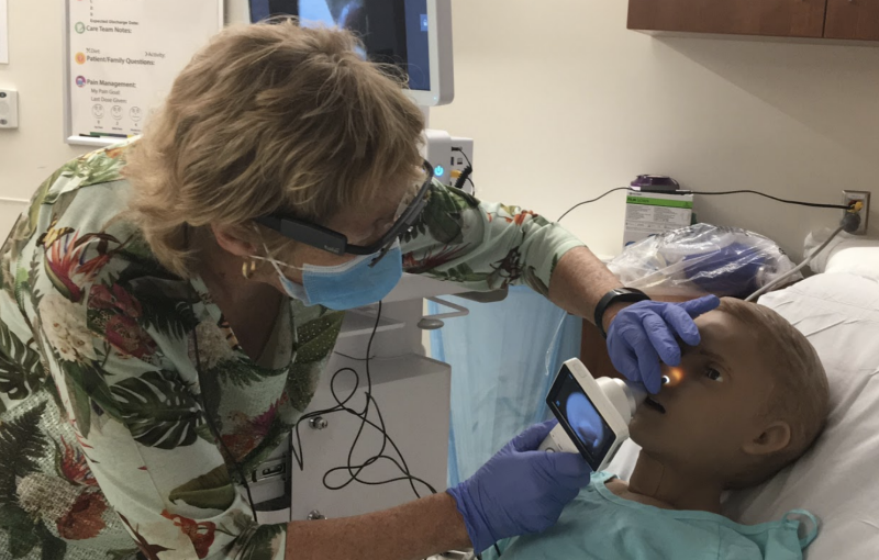 A healthcare practitioner, with her head away from the camera, is using a peripheral device from a telemedicine cart to look into the nose of a child-size mannequin on a hospital bed