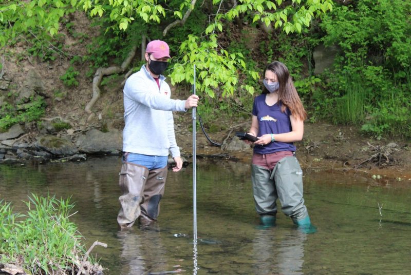 A man and a woman stand in a shallow creek. The man holds a pole upright while the woman holds and looks at a device connected to the pole by a cord.