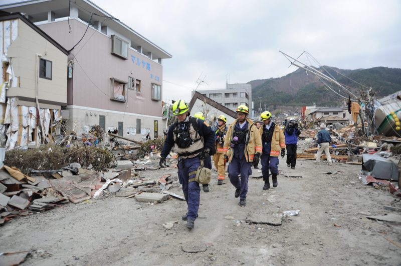 Five men in hazard gear and neon helmets walking amongst the rubble and destruction in downtown Ofunato, a coastal city in Japan.