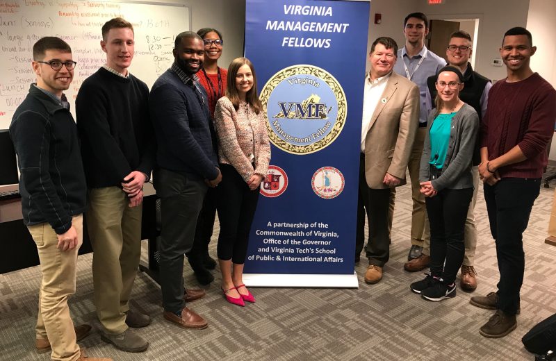 Several young adults pose in a classroom around a banner that reads "Virginia Management Fellows"