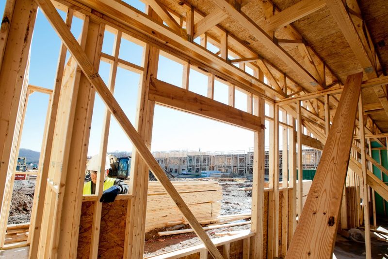 A structure under construction (framing), looking from inside the structure out. A person works on the outside of the structure, and there is a large row of units in framing in the background.