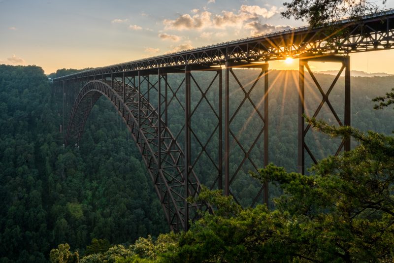 The bright, yellow sun sets over the Appalachian mountain ridge with the New River Gorge Bridge at the forefront of the image. The bridge is surrounded by lush, green trees and comes across the image, connecting two mountain sides over a river below. The bridge is rusted and dark brown with a straight section at its top and a metal arch at its bottom, connected to each other by steel beams.